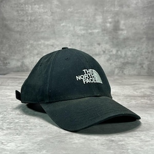 May include: A black baseball cap featuring a structured six-panel design with a curved brim. The front panel displays the white embroidered text "THE NORTH FACE" accompanied by a stylized mountain logo graphic above the brand name.