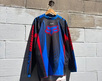 May include: A long-sleeved jersey featuring a black, blue and red colour scheme with a prominent red Fox head logo on the chest. The garment includes blue and red graphic accents along the sleeves and torso, with the word Fox printed in white.