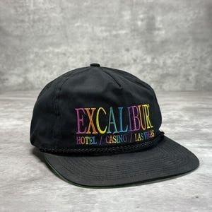 Könnte beinhalten: Schwarze Baseballkappe mit dem Wort "EXCALIBUR" in Regenbogenfarben gestickt. Darunter steht der Text "HOTEL / CASINO / LAS VEGAS". Die Kappe hat einen schwarzen Schirm und ein schwarzes Seildetail.