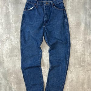 May include: A pair of classic blue denim jeans. The jeans have a button fly, five-pocket design, and a straight leg. The denim is a medium wash with some fading and wear. The jeans are laid flat on a neutral surface.