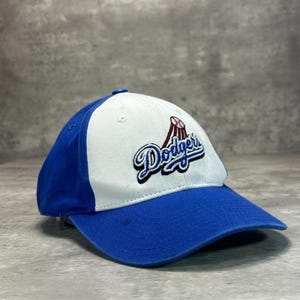 May include: A blue and white baseball cap featuring the Los Angeles Dodgers logo. The cap has a curved brim and a six-panel design. The Dodgers logo is embroidered in blue, red, and white on the front of the cap.