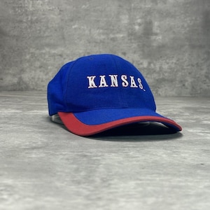 Op de afbeelding: Een koningsblauwe honkbalpet met een rode klep en het woord KANSAS in witte letters geborduurd op het voorpaneel. De pet heeft een gestructureerde kroon en een gebogen klep, uitgevoerd in een klassiek ontwerp met zes panelen.