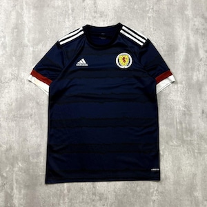 Pode incluir: Camisola de futebol Adidas azul marinho com riscas brancas nos ombros e mangas. A camisola apresenta o brasão da seleção escocesa e o logótipo da Adidas. Gola redonda e mangas curtas.