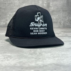 May include: Black Snap-on trucker hat with a white logo and text. The logo features tools and the text reads "Snap-on" above the names "KEVIN CIRRITO, BOB BEST, SEAN MEIERER". The hat has a black mesh back and a curved brim.