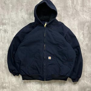 Könnte beinhalten: Dunkelmarineblaue Kapuzenjacke mit durchgehendem Reißverschluss und einer Vordertasche. Die Jacke hat schwarze gerippte Bündchen und einen Saum. Ein kleines quadratisches Logo ist vorne sichtbar. Die Jacke ist aus strapazierfähigem Stoff.