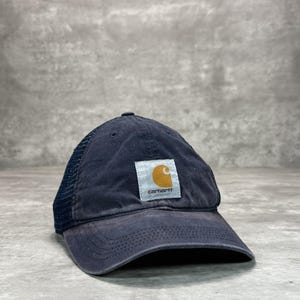 May include: Navy blue Carhartt baseball cap with a mesh back. The cap features a curved brim and a square patch with the Carhartt logo in white and gold. The hat is made of a durable material and is suitable for outdoor activities.