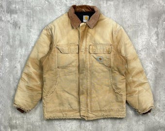 Puede incluir: Chaqueta de trabajo color beige con cuello de pana marrón. La chaqueta tiene dos bolsillos en el pecho y dos bolsillos inferiores. El logotipo de Carhartt es visible en uno de los bolsillos del pecho. La chaqueta parece desgastada.