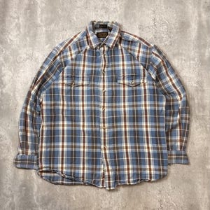 May include: A long-sleeved plaid shirt in blue, brown, and white. The shirt has a button-down front, two chest pockets, and a collared neckline. The fabric appears to be a medium-weight cotton or blend, suitable for casual wear.