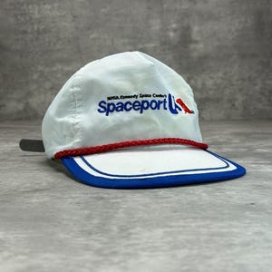 May include: A white baseball cap featuring a blue and red embroidered logo that reads NASA Kennedy Space Center's Spaceport. The cap includes a bright red braided cord across the base of the crown and a blue brim with white trim.