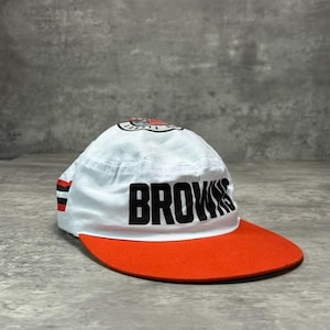 May include: A white baseball cap with a flat orange brim, featuring the word BROWNS in bold black lettering on the front. The cap includes a small circular logo on the top crown and horizontal black and orange stripes on the side panel.