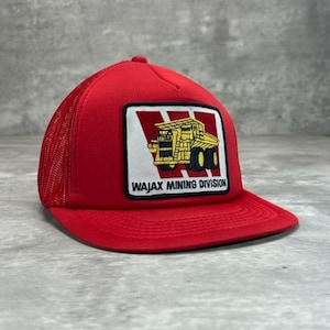 May include: Red trucker hat with a mesh back and a curved brim. The front features a rectangular patch with a yellow and black mining truck graphic and the text "WAJAX MINING DIVISION".