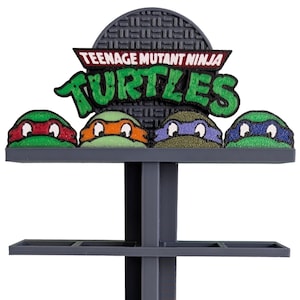 Op de afbeelding: Een grijze kunststof displaystandaard met vier niveaus, elk voorzien van twee vierkante vakken. De bovenkant bevat het Teenage Mutant Ninja Turtles-logo in groen en rood, met vier schildpadmaskers in de kleuren rood, oranje, paars en blauw.
