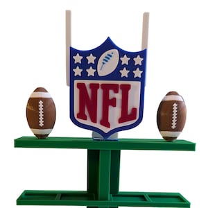 May include: A green, multi-tiered display stand featuring the NFL logo and two miniature American footballs. The logo is blue, white, and red. The stand has multiple shelves for displaying items. The footballs are brown with white laces.