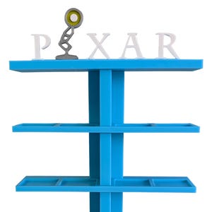 Può includere: Un espositore blu brillante a cinque livelli con un supporto verticale centrale. Il ripiano superiore presenta lettere bianche che compongono la parola PIXAR, con una lampada da scrivania articolata grigia che sostituisce la I, dotata di un elemento luminoso circolare giallo.