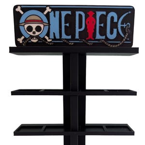 Peut inclure: Un présentoir noir à quatre niveaux avec une enseigne supérieure affichant le logo One Piece, comprenant un crâne portant un chapeau de paille, des lettres bleues et la silhouette rouge d'un personnage sur un fond sombre.