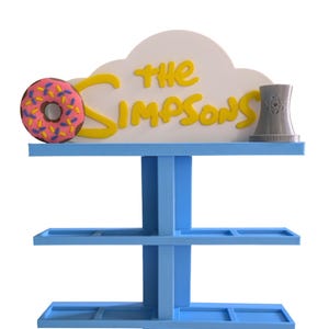 Może przedstawiać: Niebieska półka ekspozycyjna z żółtym napisem "The Simpsons", różowym pączkiem z posypką i szarą elektrownią jądrową. Półka ma kilka poziomów do eksponowania przedmiotów.