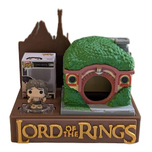 Peut inclure: Un présentoir marron du Seigneur des Anneaux avec une miniature de trou de Hobbit, une figurine et une boîte. Le trou de Hobbit a un toit vert et une porte ronde. La boîte contient une figurine de Frodon. Les mots "LORD OF THE RINGS" sont en lettres dorées.