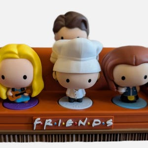 Könnte beinhalten: Ein orangefarbenes Miniatursofa mit dem Friends-Logo auf der Vorderseite, besetzt mit drei stilisierten Figuren. Die Figuren umfassen eine blonde Figur mit Gitarre, eine Figur mit weißer Kochmütze und eine Figur mit braunem Haar.