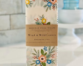 May include: Cream-coloured beeswax food wraps featuring a floral design. The wraps display a pattern of colourful flowers and green leaves. The label reads "Wind & Wildflowers" and "Bread Wrap XXL (43x53cm)".