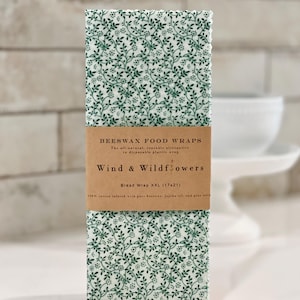 May include: A stack of beeswax food wraps with a green and white floral design. The wraps are labelled "Wind & Wildflowers" and are for bread, measuring 43x53 cm. The label states they are an all-natural, reusable alternative to disposable plastic wrap.