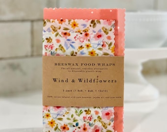 May include: A pack of three reusable beeswax food wraps with floral designs and a coral pink edge. The wraps are branded "Wind & Wildflowers" and made from cotton infused with beeswax, jojoba oil, and pine resin. Sizes are 20x20, 23x23, and 25x25 cm.