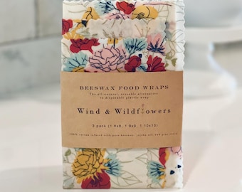 May include: A pack of three beeswax food wraps featuring a floral design in red, yellow, and blue. The wraps are labelled "Wind & Wildflowers" and come in sizes 20x20, 23x23, and 25x25 cm. The packaging states they are a reusable alternative to disposable plastic wrap.