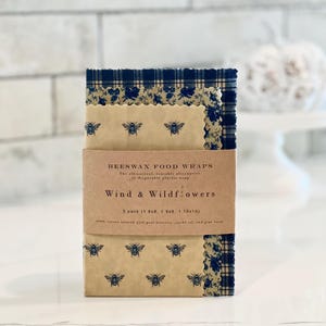 May include: A set of three beeswax food wraps in a brown cardboard box. The wraps are printed with a blue and white floral pattern and have a bee design. The box says "Beeswax Food Wraps" and "Wind & Wildflowers". The box also says "3 pack (1 8x8, 1 9x9, 1 10x10)".