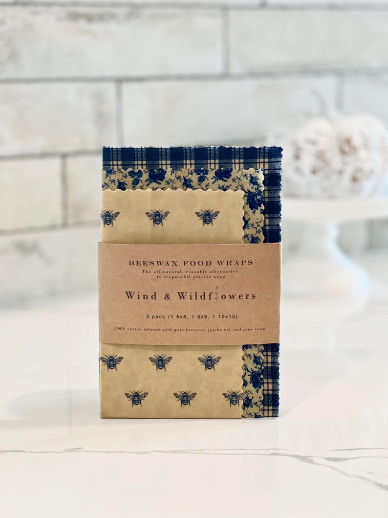 May include: A set of three beeswax food wraps in a brown cardboard box. The wraps are printed with a blue and white floral pattern and have a bee design. The box says "Beeswax Food Wraps" and "Wind & Wildflowers". The box also says "3 pack (1 8x8, 1 9x9, 1 10x10)".