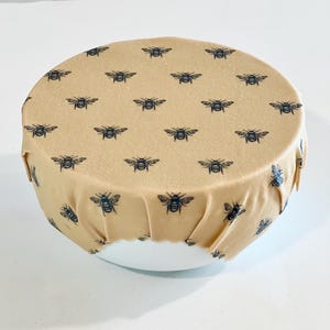 May include: A beige reusable food wrap with a black bee pattern. The wrap is covering a white bowl.