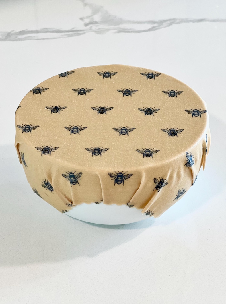May include: A beige reusable food wrap with a black bee pattern. The wrap is covering a white bowl.