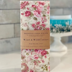 Puede incluir: Un envoltorio rectangular de cera de abeja para alimentos con un estampado floral en rosa y rojo sobre un fondo crema. La etiqueta dice "Wind & Wildflowers" y "Bread Wrap XXL (43x53cm)". Alternativa reutilizable.