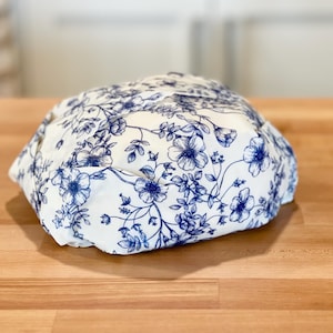 May include: A white fabric item with a blue floral pattern, resting on a wooden surface. The floral design features detailed blue flowers and leaves, creating a classic and elegant look. The item appears to be a hat or a similar head covering.