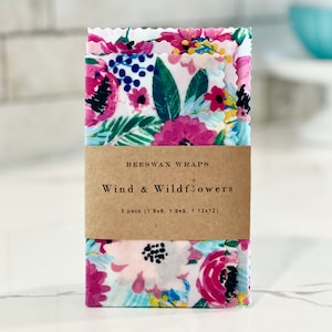 May include: Three beeswax food wraps with a floral print in shades of pink, blue, and green. The wraps are labeled "Beeswax Wraps Wind & Wildflowers 3 pack (1 8x8, 1 9x9, 1 12x12)"