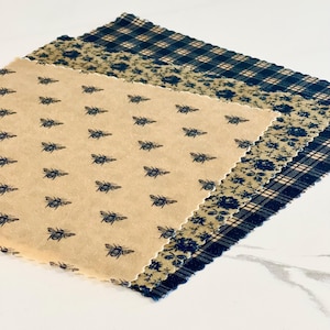 May include: Three reusable food wraps with different patterns. The top wrap is a black and white plaid pattern. The middle wrap is a floral pattern with a beige background. The bottom wrap has a beige background with a repeating pattern of bees.