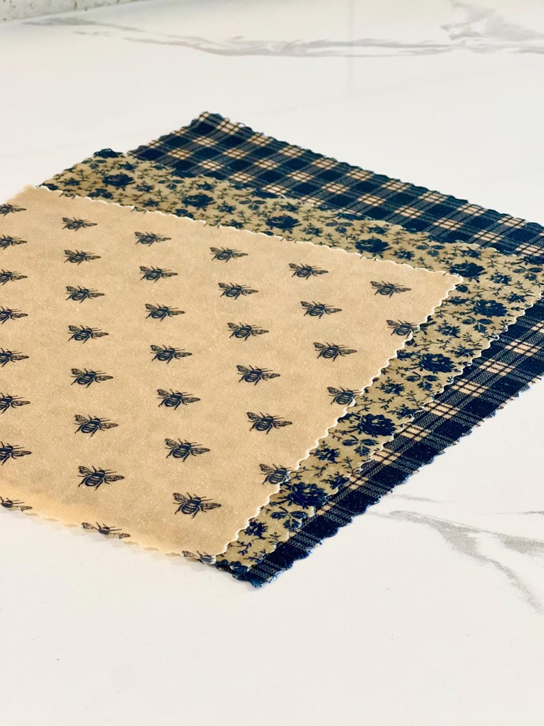 May include: Three reusable food wraps with different patterns. The top wrap is a black and white plaid pattern. The middle wrap is a floral pattern with a beige background. The bottom wrap has a beige background with a repeating pattern of bees.
