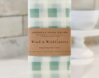 May include: A green and white gingham beeswax food wrap labelled "Beeswax Food Wraps" and "Wind & Wildflowers". The label also states "Bread Wrap XXL (17x21)" and "100% cotton infused with pure beeswax, jojoba oil, and pine resin".