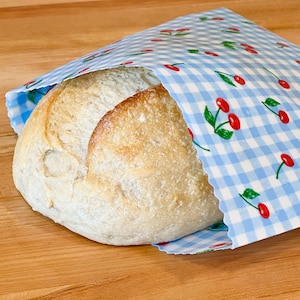 May include: A loaf of bread peeks out from a blue and white gingham bag decorated with red cherries and green leaves. The bread has a golden-brown crust and a light-colored interior. The bag is open, revealing the bread.