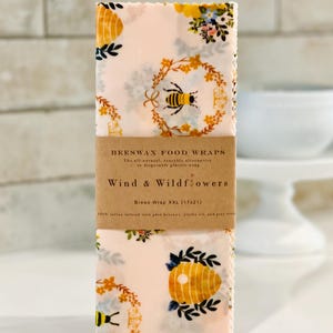 Puede incluir: Envolturas de alimentos de cera de abeja con un patrón floral rosa y amarillo con un diseño de abeja y panal. La etiqueta dice "Beeswax Food Wraps, The all-natural, reusable alternative to disposable plastic wrap, Wind & Wildflowers, Bread Wrap XXL (17x21)"