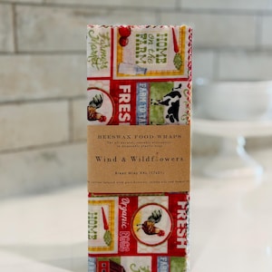 May include: A rectangular beeswax food wrap with a farm-themed print featuring roosters, cows, and vegetables in red, yellow, and green. A brown paper band labeled Wind & Wildflowers Bread Wrap XXL (17x21) is wrapped around the center of the fabric.