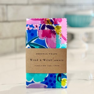 May include: Three beeswax food wraps with a floral pattern in shades of pink, blue, and green. The wraps are packaged together with a brown paper label that reads "BEESWAX WRAPS Wind & Wildflowers 3 pack (1 8x8, 1 9x9, 1 10x10)"