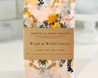 May include: A pack of beeswax food wraps featuring a floral and bee pattern. The wraps are pink and white, adorned with bees, wildflowers, and honeycombs. The label says "Wind & Wildflowers" and lists sizes as 20x20, 23x23, and 25x25 cm.