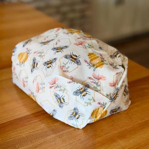 May include: A rectangular fabric bread warmer featuring a cream-colored background with a repeating pattern of honeybees, yellow honeycomb outlines, and small pink floral sprigs. The fabric is folded into a structured shape with visible seams.