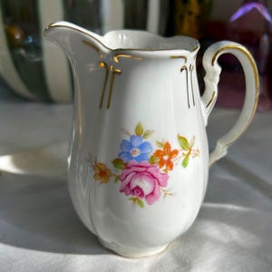 May include: A white porcelain creamer with a gold trim and floral design. The flowers are pink, blue, and orange.