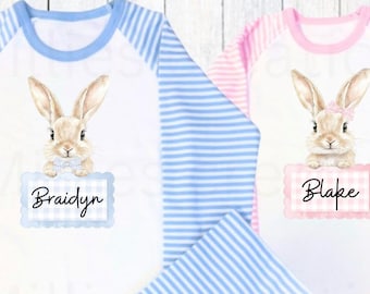 May include: Two long-sleeved pajamas with a white background and blue and pink stripes on the sleeves. The pajamas feature a brown bunny rabbit holding a sign with a name on it. The left pajama has a blue sign with the name "Braidyn" and the right pajama has a pink sign with the name "Blake".