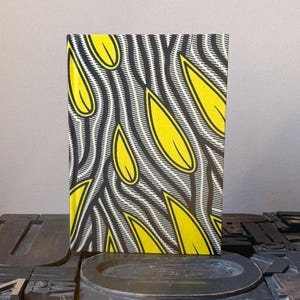 May include: A rectangular book with a vibrant cover design. The cover features a pattern of yellow leaf shapes against a backdrop of black and white striped lines. The book is displayed on a dark surface.