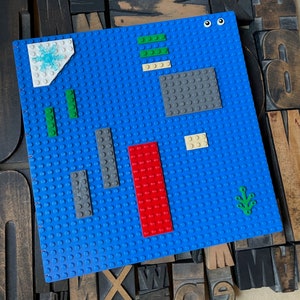 Large coptic bound blank book made from blue LEGO baseplates that measure 10 inches square. The book is decorated with 25 LEGO pieces in various colors. The paper is 70 pound paper suitable for drawing. The book is shown standing from the back.