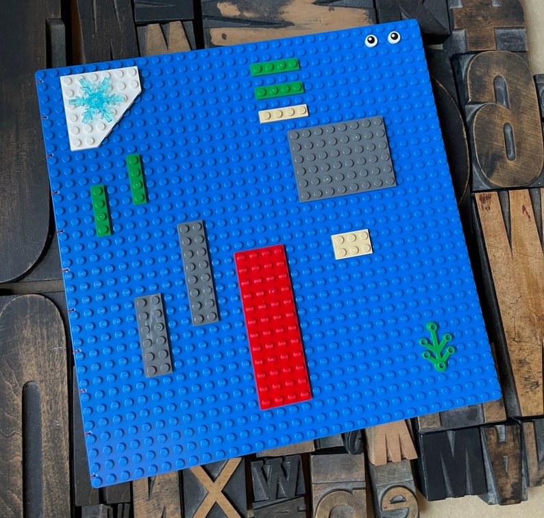 Large coptic bound blank book made from blue LEGO baseplates that measure 10 inches square. The book is decorated with 25 LEGO pieces in various colors. The paper is 70 pound paper suitable for drawing. The book is shown standing from the back.