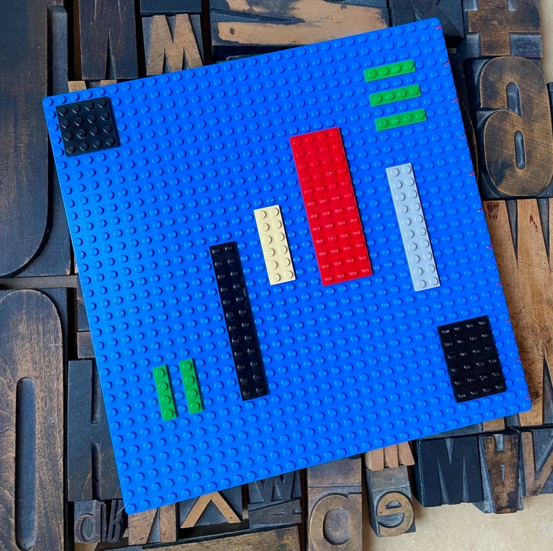 Large coptic bound blank book made from blue LEGO baseplates that measure 10 inches square. The book is decorated with 25 LEGO pieces in various colors. The paper is 70 pound paper suitable for drawing. The book is shown laying flat from the front.