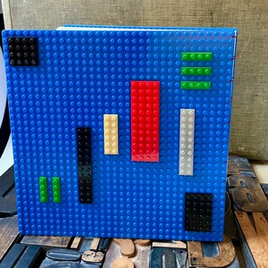 Large coptic bound blank book made from blue LEGO baseplates that measure 10 inches square. The book is decorated with 25 LEGO pieces in various colors. The paper is 70 pound paper suitable for drawing. The book is shown standing from the back.