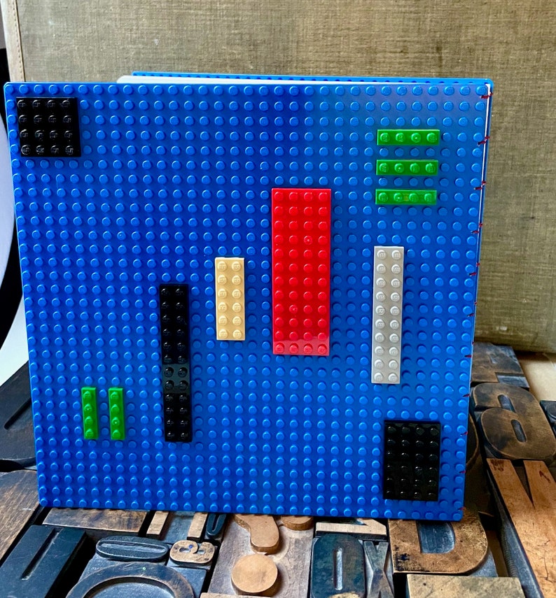 Large coptic bound blank book made from blue LEGO baseplates that measure 10 inches square. The book is decorated with 25 LEGO pieces in various colors. The paper is 70 pound paper suitable for drawing. The book is shown standing from the back.